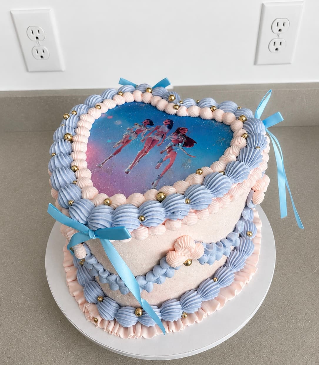 Two-tier heart-shaped birthday cake with blue and blush pink shell piping, gold pearl accents, blue ribbon bows, and edible character image topper